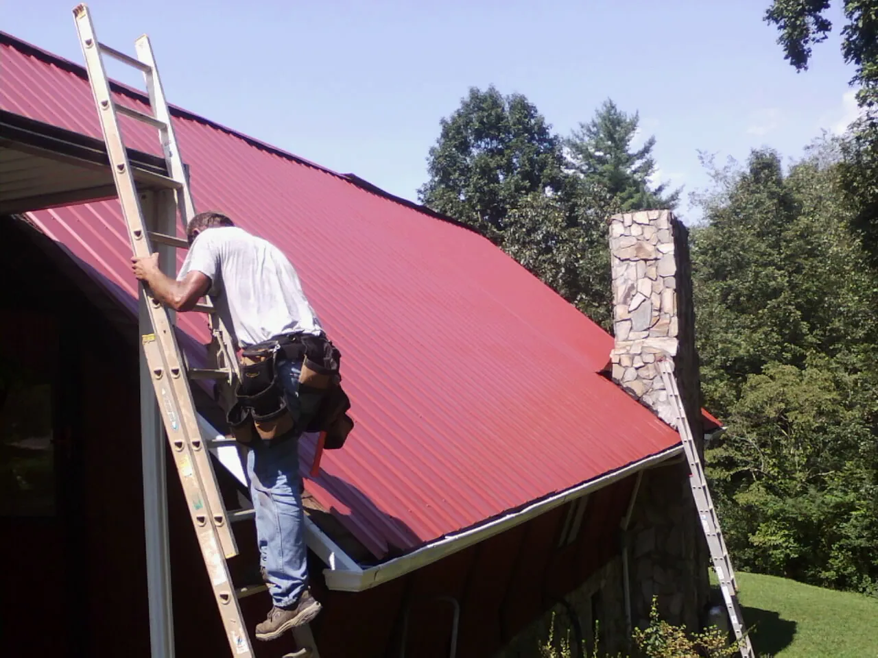 Roofer on a ladder beside a red metal roof and stone chimney — Soffit & Fascia Repair by Roofing Brothers in Whiteside
